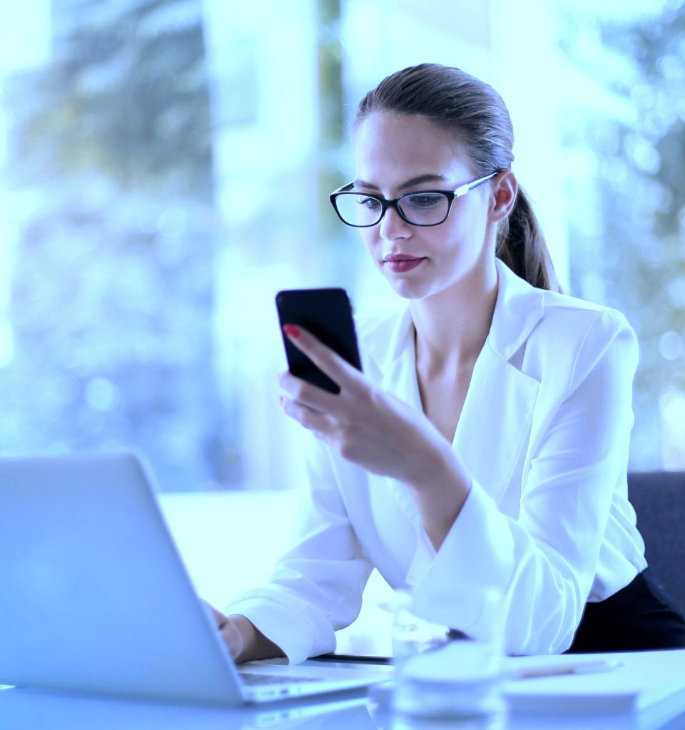 A woman wearing blue light glasses while sitting at a desk with her phone in hand and laptop open in front of her