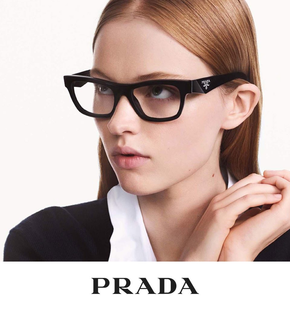 A woman wearing thick, black, rectangular Prada eyeglasses featuring a small silver brand logo on the arm