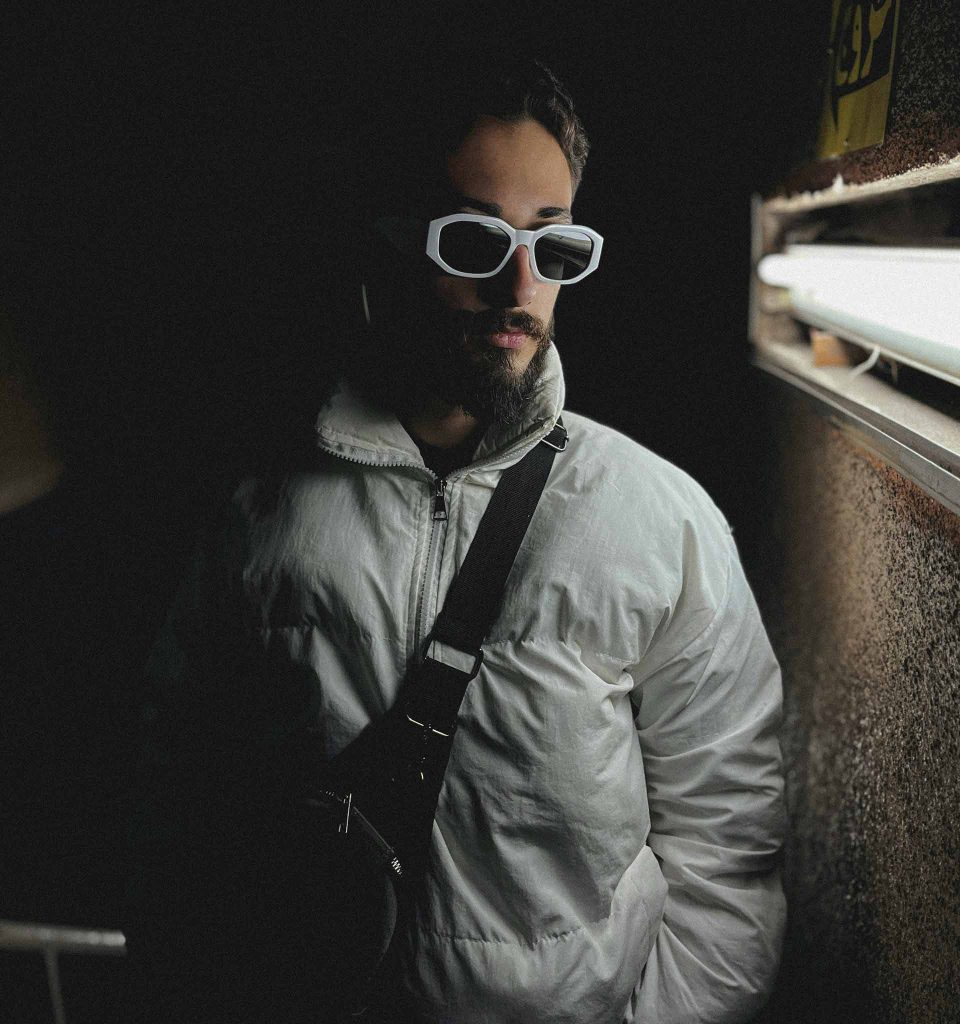 Man wearing grey puffer coat, black crossbody bag and white sunglasses