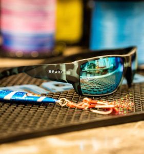 Bajío sunglasses with blue polarized lenses and camo frames, placed on a textured surface with a fishing lure nearby
