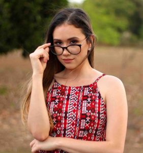 A young woman with long brown hair adjusting the frames of her round black glasses