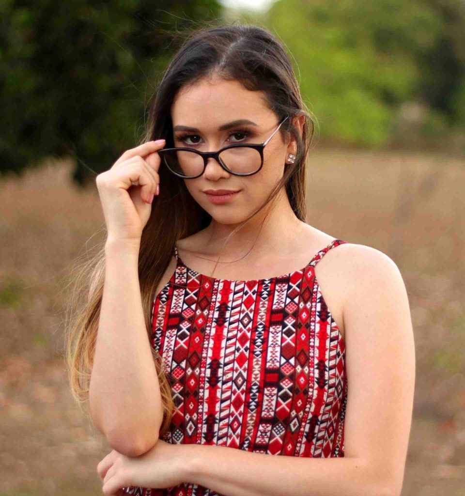 A young woman with long brown hair adjusting the frames of her round black glasses