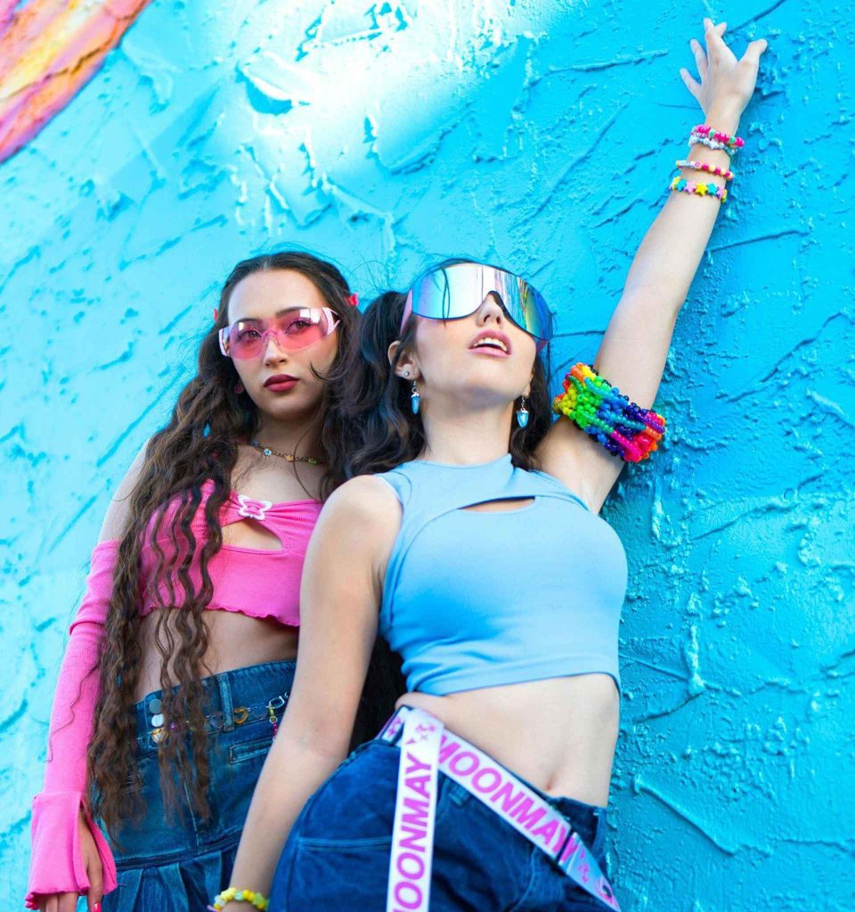 Two young women in Y2K sunglasses posing against a textured blue wall
