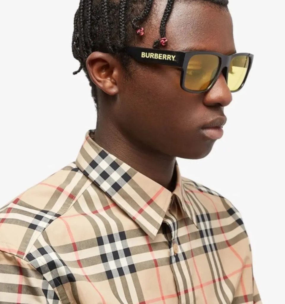 A model wearing Burberry Knight sunglasses with thick black frames, yellow lenses, and arms embellished with the brand name