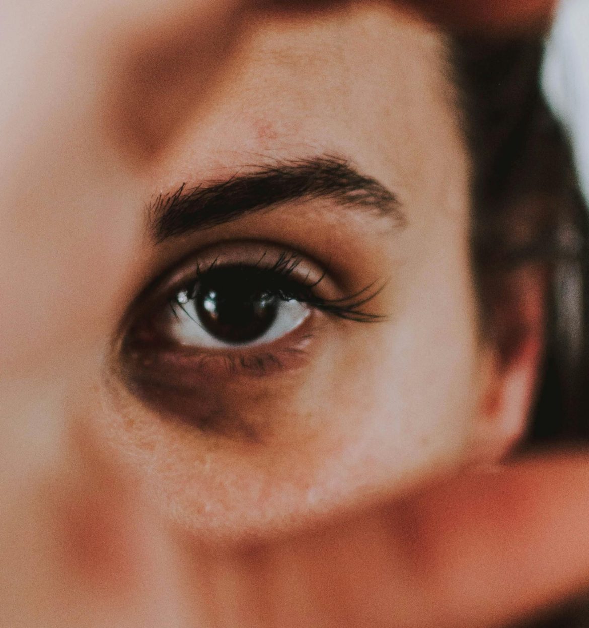 A close-up photo of a deep brown eye focused on the camera lens through the space between the person’s thumb and index finger