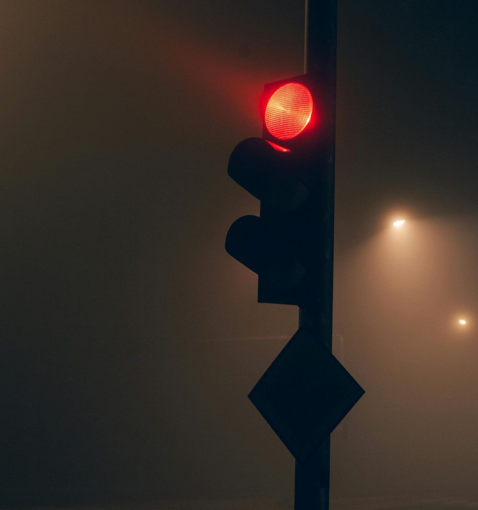 A traffic light, on a dark, foggy road, signaling red with a traffic sign barely visible below