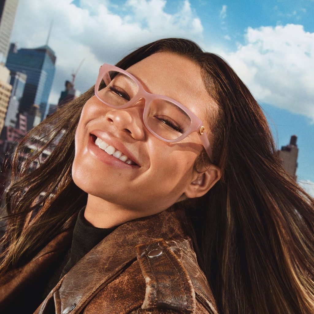 A smiling woman wearing baby pink Coach eyeglass frames and a brown leather jacket while standing in front of a cityscape