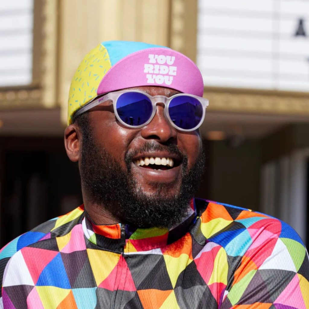 A smiling man wearing a brightly colored shirt and hat with translucent white, round Goodr sunglasses
