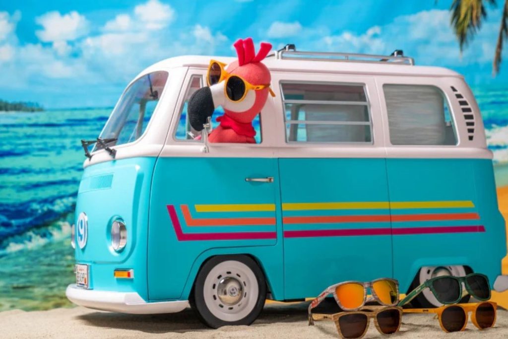 The Goodr pink flamingo driving a blue Volkswagen bus as sunglasses sit in the foreground
