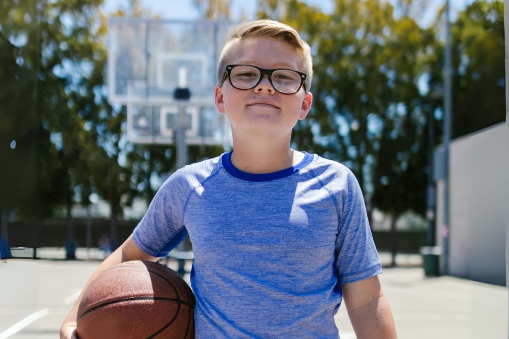 A boy on a basketball court, holding a ball and wearing polycarbonate eyeglasses