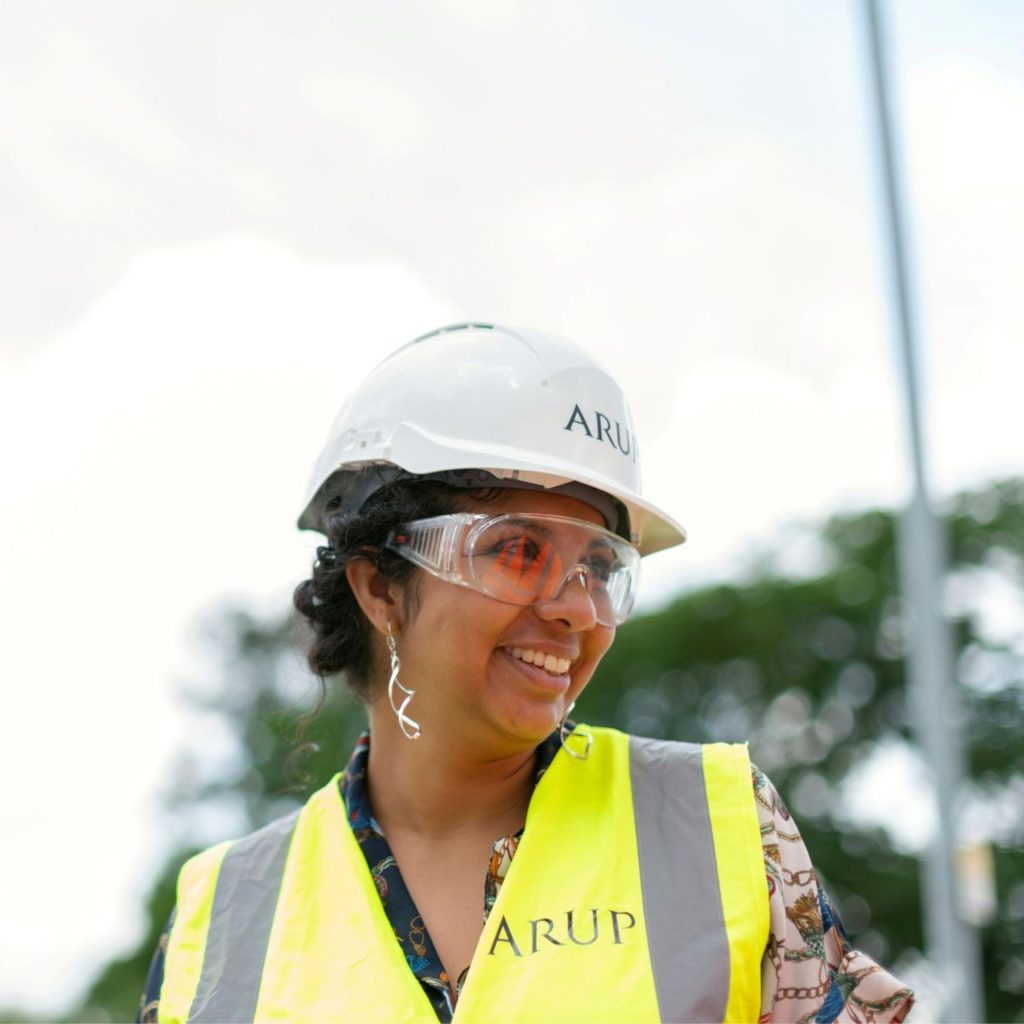 A woman in a high-vis vest, hard hat, and protective goggles smiles at something off-camera