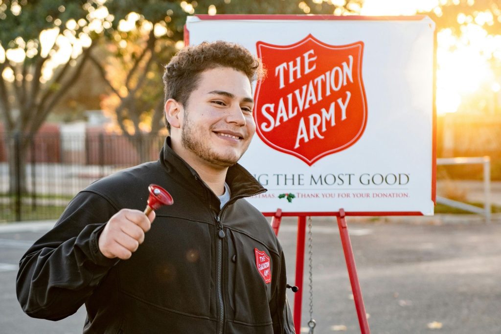 A Salvation Army volunteer ringing a red bell as he stands next to a donation sign