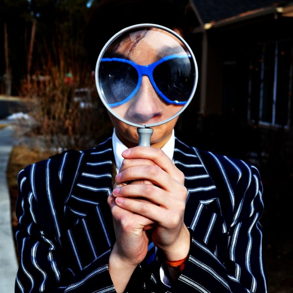 A man in a striped suit and sunglasses looking through a large magnifying glass