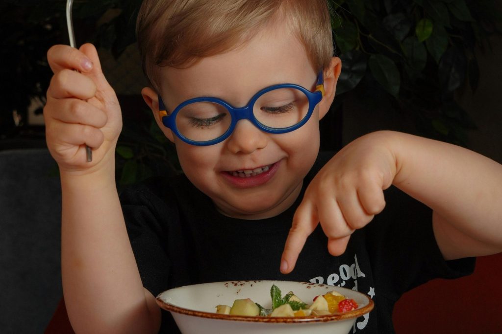 A child wearing round blue eyeglasses smiles as he plays with food in a bowl