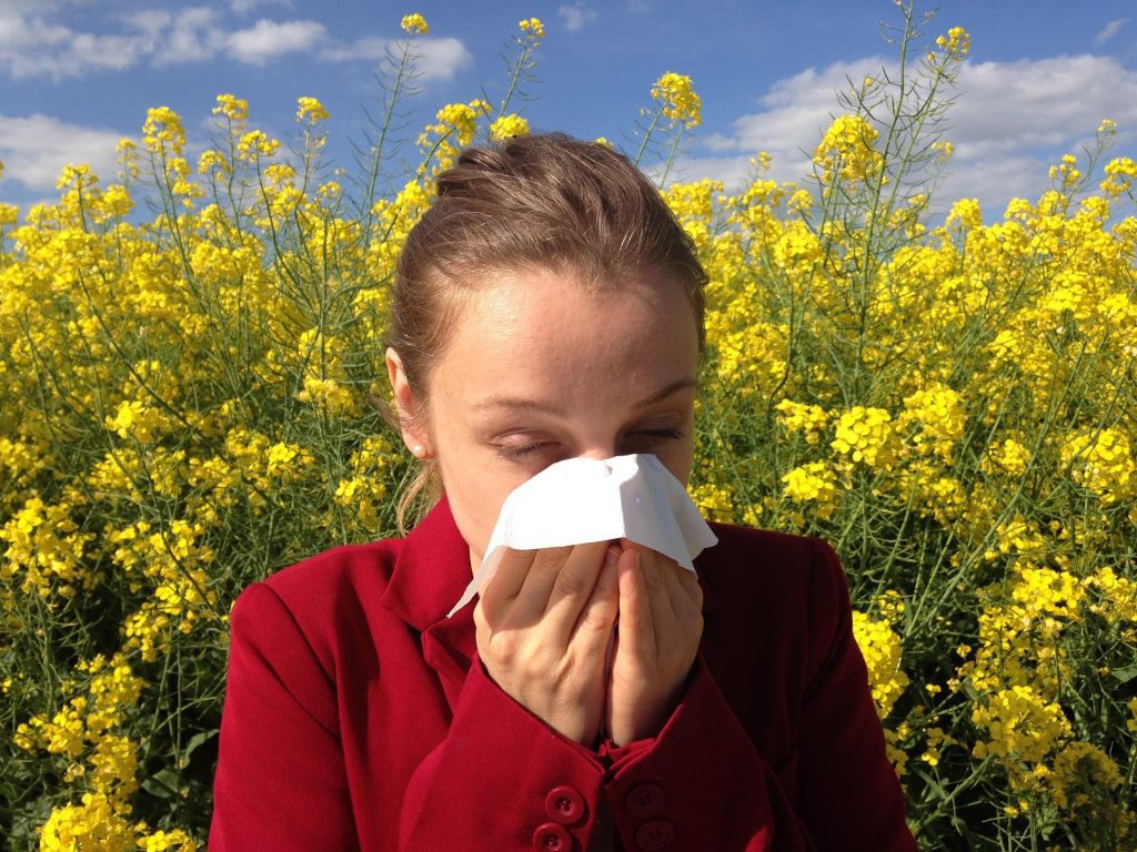 A woman in a field of flowers experiencing symptoms of allergies and holding a tissue to her nose