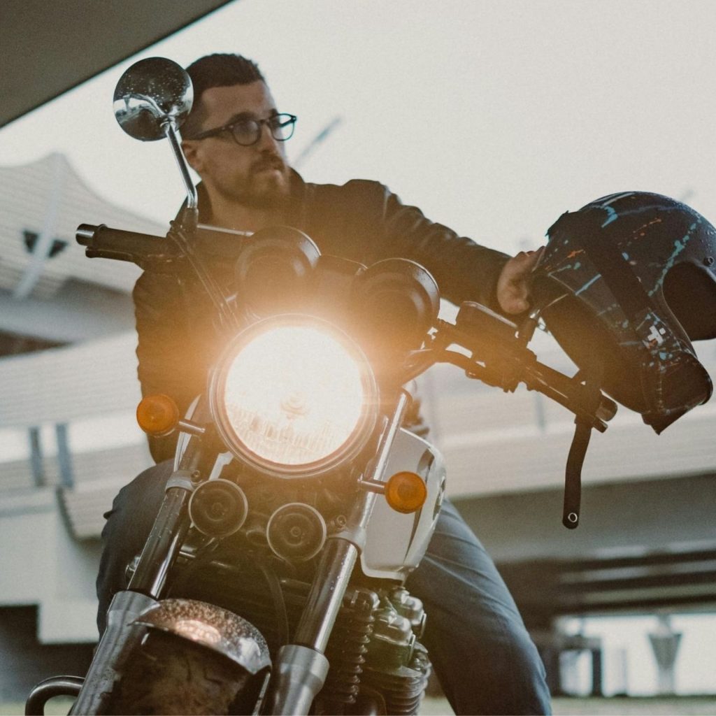 Man with glasses sits on a motorcycle while holding his helmet