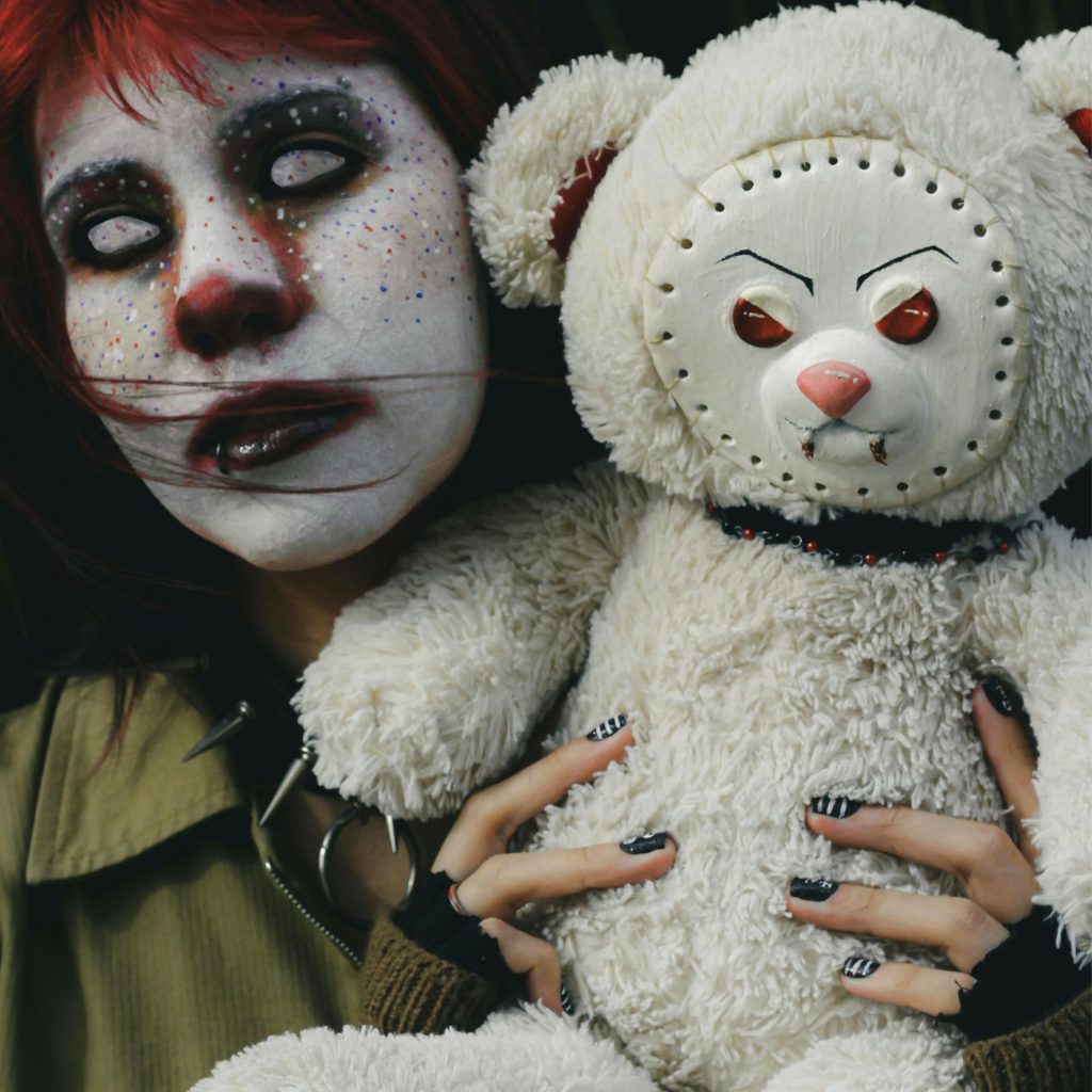 Woman in full face white makeup and white contacts, holding a scary doll