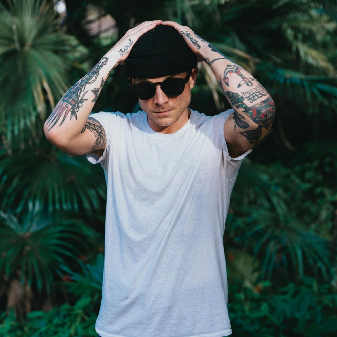 Man with tattooed arms, wearing white shirt, jeans, and oversized sunglasses