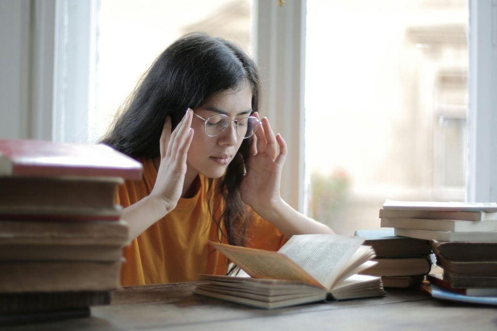 A woman in glasses rubs her temples; she is surrounded by books