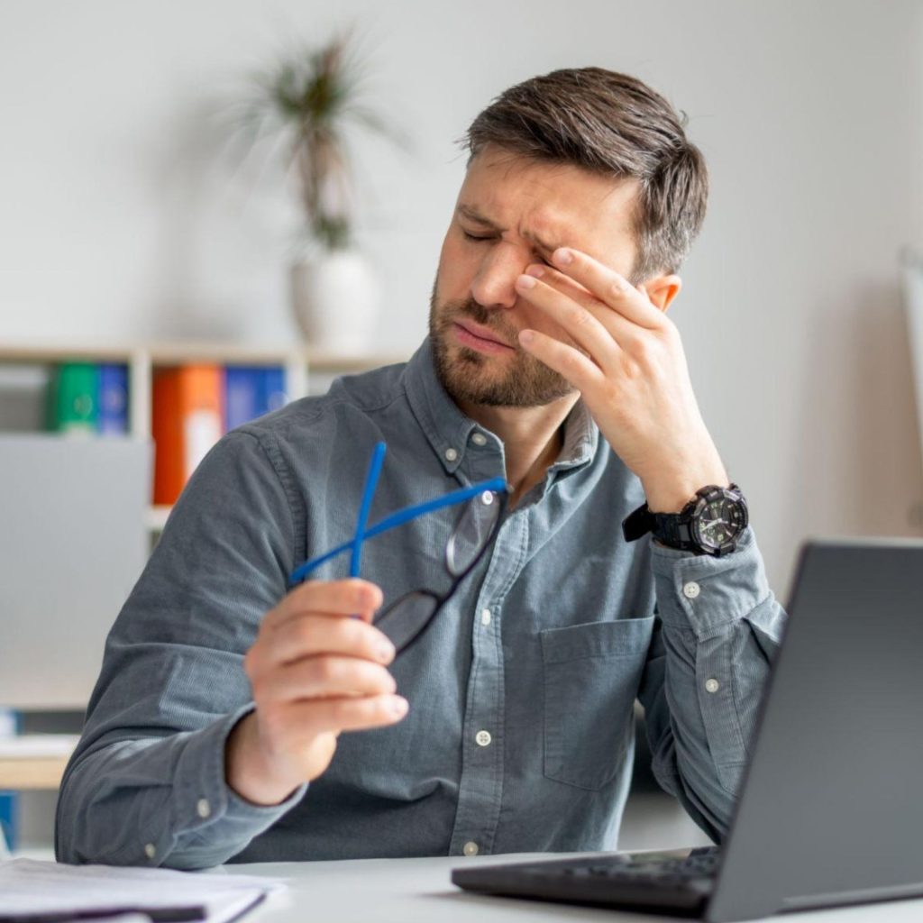 Man holds glasses and rubs his eye in front of a laptop