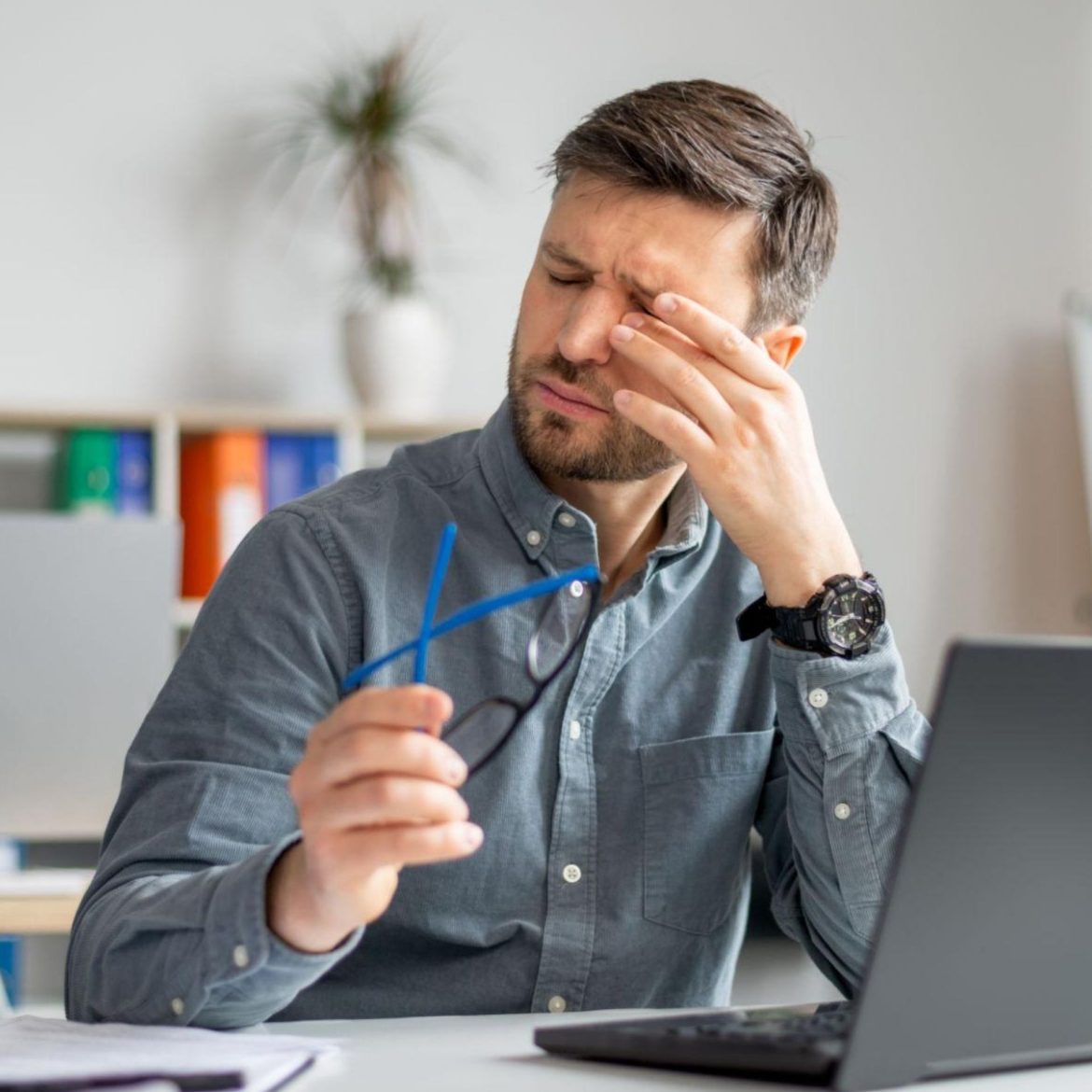 Man holds glasses and rubs his eye in front of a laptop
